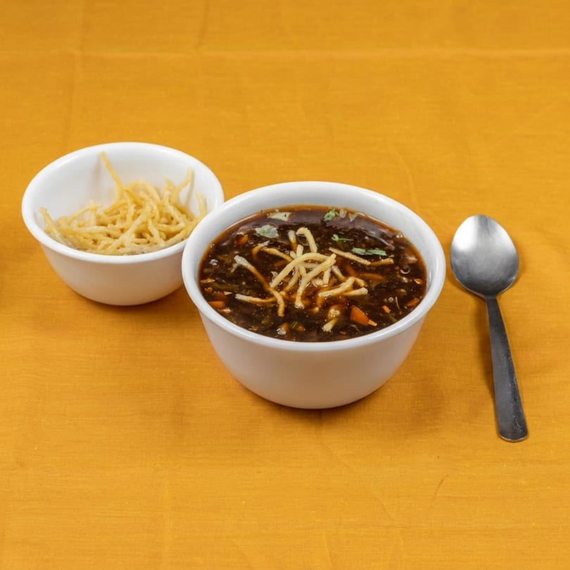 Hot and sour soup with crispy noodles in white bowls, alongside a spoon on a yellow tablecloth.
