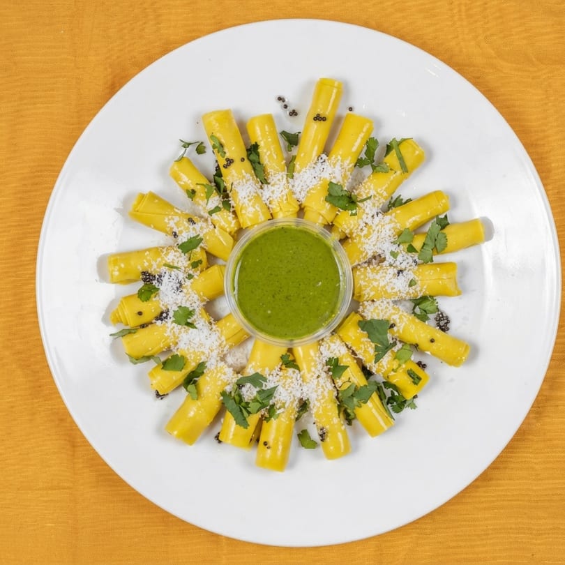 Yellow khandvi rolls arranged in a circle with green chutney, grated coconut, and cilantro.