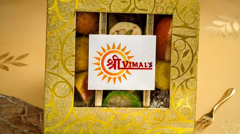 A gold box of assorted Indian sweets with a "Shree Vimal's" logo, next to a gold fork.