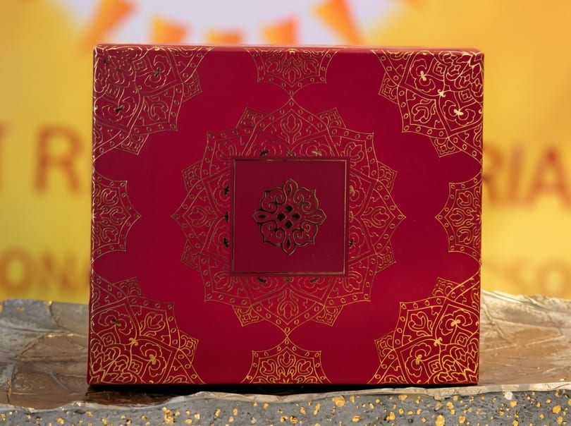 Red box with intricate gold patterns, featuring a central medallion design, on a textured gray surface.