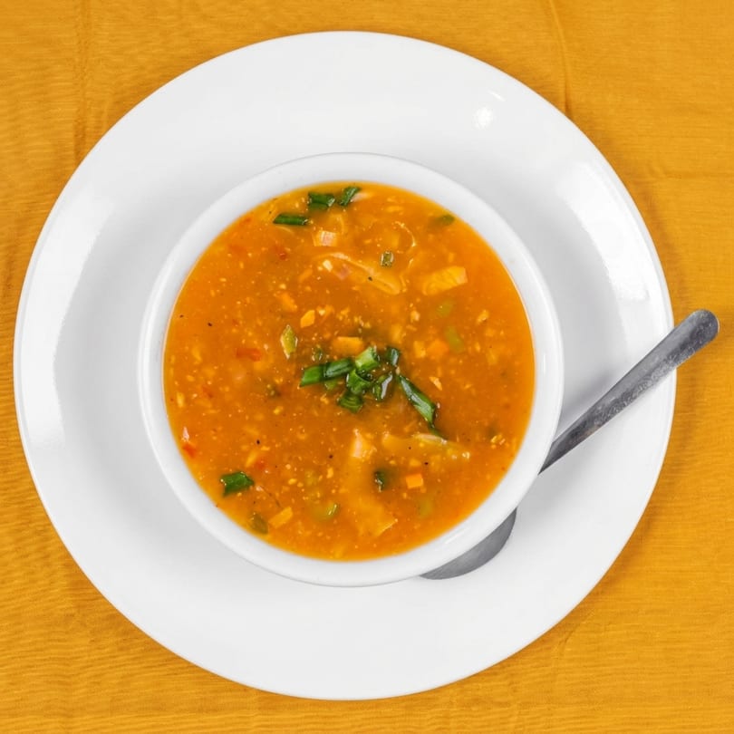 Overhead view of a bowl of orange soup with vegetables and green onions on a white plate.